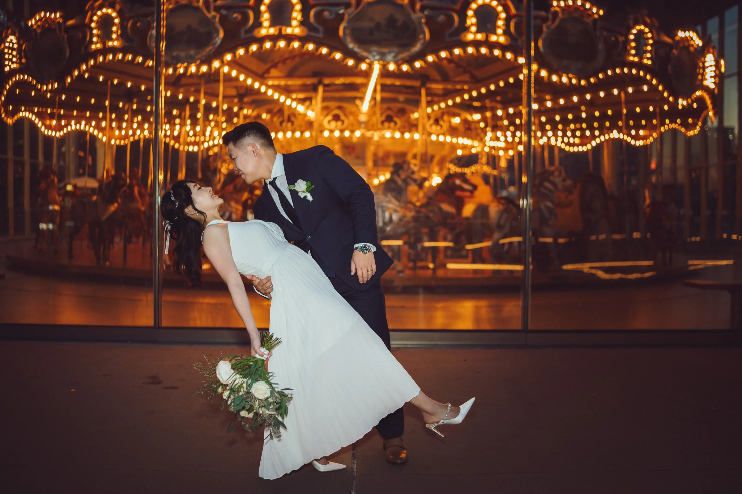 Groom lifting the bride in front of a carousel glowing with golden lights
