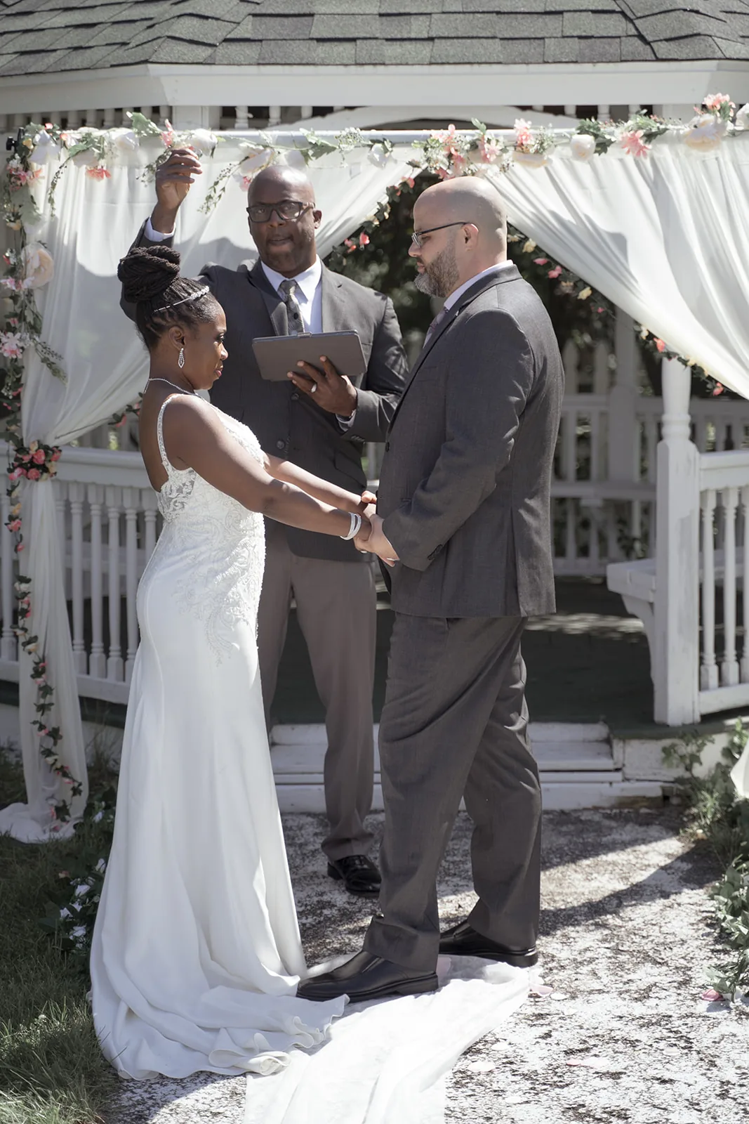 Bride and groom holding hands during a wedding ceremony beside a flower-decorated garden gazebo