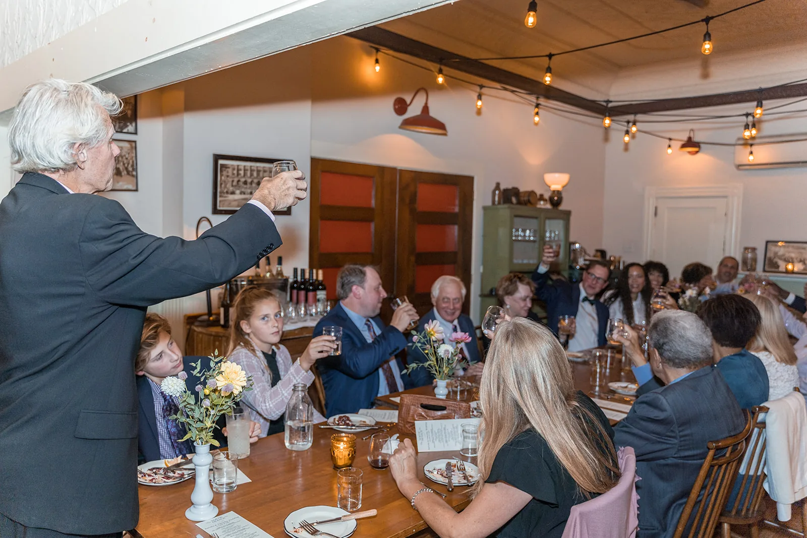 Wedding guests raising their glasses during a reception toast beneath string lights