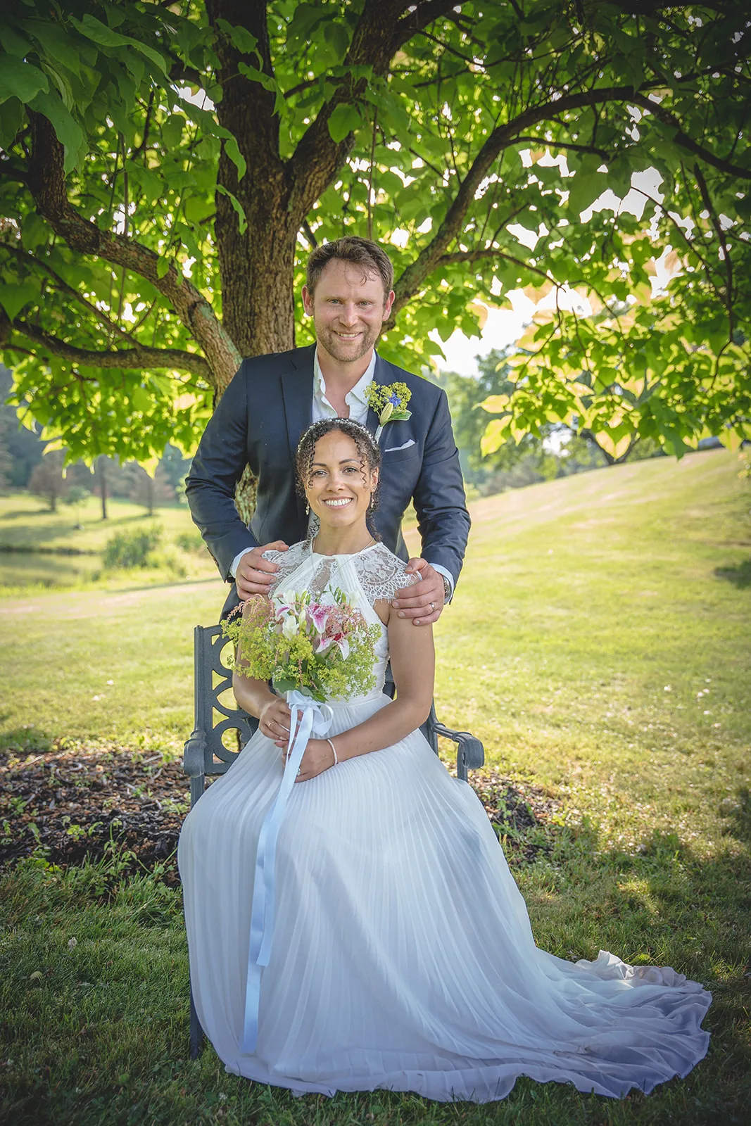 Seated bride holding her bouquet with her groom behind her in sunlit woods